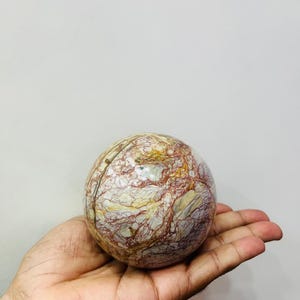 Può includere: Una sfera di pietra lucida con un motivo marmorizzato nei toni del rosso, crema e oro. La sfera è tenuta in una mano aperta su uno sfondo bianco. È un oggetto decorativo.