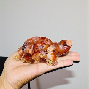 May include: A decorative turtle figurine crafted from translucent, reddish-brown and orange stones. The turtle's shell and head are detailed with intricate patterns, and it has gold-colored accents around the edges. The figurine is held in a hand.