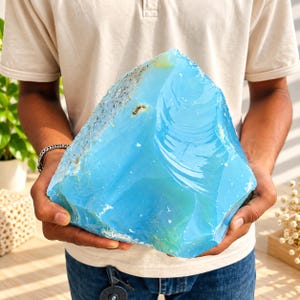 Può includere: Un grande esemplare di minerale grezzo color azzurro cielo, traslucido, con una consistenza liscia simile al vetro su un lato e una crosta ruvida, maculata di bianco e grigio lungo il bordo superiore, tenuto saldamente per mostrarne la forma.