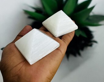 Puede incluir: Dos pirámides de selenita blanca se sostienen en una mano, mostrando su forma geométrica y textura natural. Las pirámides se colocan sobre un fondo de follaje verde, resaltando su diseño limpio y minimalista. Ideal para la decoración.