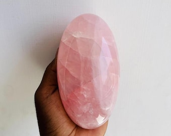 May include: A polished, oval-shaped rose quartz crystal with a smooth, translucent surface. The stone displays a soft, pale pink hue with natural internal veining and crystalline structures visible throughout its rounded, symmetrical form.