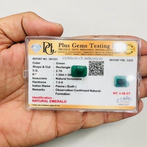 Op de afbeelding: Een rechthoekige natuurlijke smaragd met een gewicht van 4,48 CT, gepresenteerd in een doorzichtige plastic houder met een certificaat van echtheid van Plus Gems Testing Lab. Het document vermeldt specificaties zoals kleur, vorm en hardheid.
