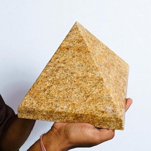 May include: A golden-brown, pyramid-shaped stone sculpture. The pyramid is held in a hand against a white background. The stone has a textured surface and a geometric shape, suitable for home decor.