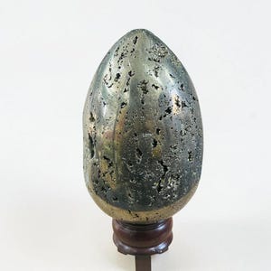 May include: A metallic, egg-shaped pyrite specimen with a rough, textured surface. The pyrite is a dark, gunmetal grey colour with flecks of gold. It is mounted on a small, dark brown wooden stand.