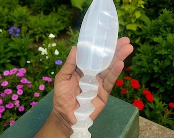 May include: A clear, selenite wand with a leaf-shaped top and a spiraling handle. The wand is held in a hand against a backdrop of green foliage and colorful flowers. The wand is approximately 8 inches long.