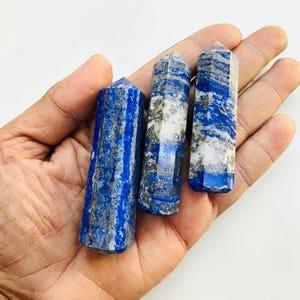 May include: Three polished lapis lazuli crystal towers. The blue stones have white and gray veining and are held in a hand. The crystals are hexagonal and have a pointed top.