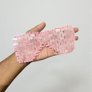 Op de afbeelding: Een roze oogmasker gemaakt van kleine, rechthoekige rozenkwartsstenen. Het masker wordt in een hand gehouden en heeft een kralenrand. De stenen zijn gerangschikt in een baksteenachtig patroon, waardoor een textuur ontstaat. Het masker is ontworpen om de ogen te bedekken.