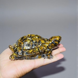 May include: A decorative turtle figurine, crafted from a mosaic of golden and brown stones. The turtle is resting on a hand, showcasing its detailed design and natural color variations. The figurine's shell and body are intricately detailed.