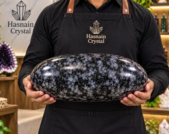 May include: A large, smooth, oval-shaped stone with a polished finish. The stone features a dark black base color speckled with numerous small, star-like white and light gray crystalline patterns distributed evenly across its entire surface.