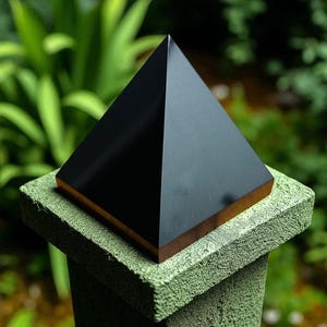 Puede incluir: Una escultura de pirámide negra con una base de color bronce se asienta sobre un poste de hormigón pintado de verde y desgastado. La superficie lisa y reflectante de la pirámide contrasta con el poste texturizado, sobre un fondo verde borroso.