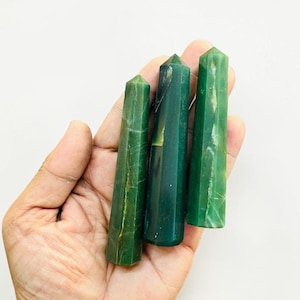 Op de afbeelding: Drie groene edelsteen obelisken van verschillende grootte worden in een hand gehouden. De obelisken hebben een gepolijste afwerking en een natuurlijk, gemarmerd uiterlijk. De edelstenen hebben een diepe, rijke groene kleur.
