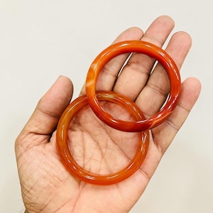 Puede incluir: Dos brazaletes de ágata de color naranja rojizo. Los brazaletes translúcidos tienen un acabado pulido, con variaciones naturales de color. Los brazaletes son redondos y parecen de tamaño mediano. Las joyas se exhiben en una mano.