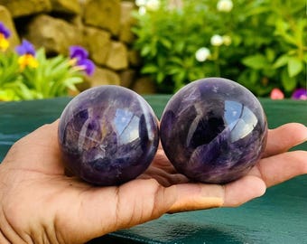 Op de afbeelding: Twee gepolijste amethistbollen, elk met een dieppaarse tint en wervelende patronen, worden in een hand gehouden. De bollen reflecteren licht, waardoor een glanzend uiterlijk ontstaat. De achtergrond toont groen en bloemen.