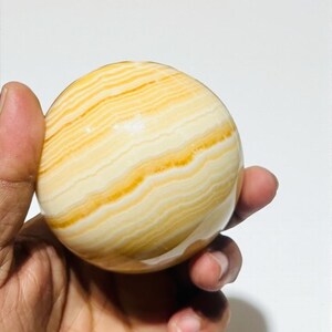 May include: A polished, spherical decorative stone with a banded pattern of creamy white and golden yellow hues. The orb is held in a hand, showcasing its smooth, rounded form. This natural stone sphere is a decorative item.