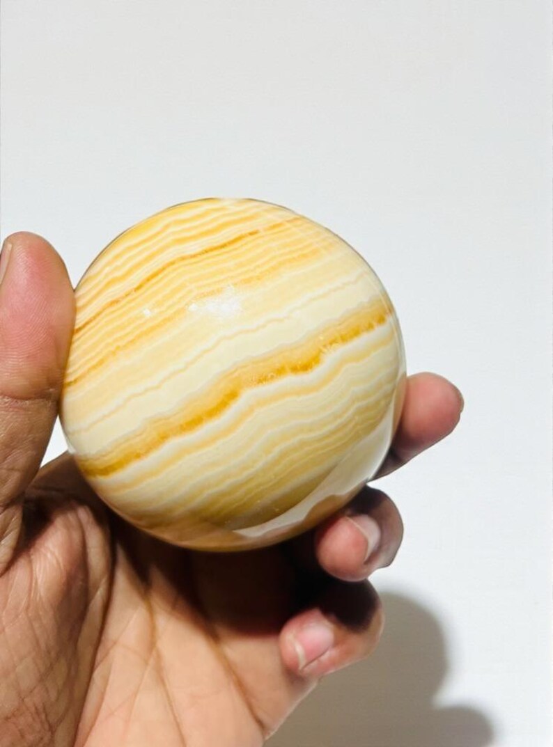 May include: A polished, spherical decorative stone with a banded pattern of creamy white and golden yellow hues. The orb is held in a hand, showcasing its smooth, rounded form. This natural stone sphere is a decorative item.