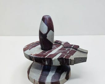 May include: A carved stone sculpture featuring a cylindrical form atop a tiered base. The sculpture is primarily gray with maroon stripes and accents. The top portion has a rounded, oval shape. The overall design suggests a spiritual or decorative object.