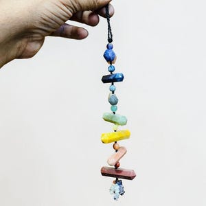 Könnte beinhalten: Ein farbenfrohes Chakra-Stein-Hängeornament. Das Ornament besteht aus polierten Edelsteinen in Blau, Grün, Gelb und Rot, die an einer schwarzen Schnur aufgereiht sind. Die Steine haben unterschiedliche Formen und Größen.