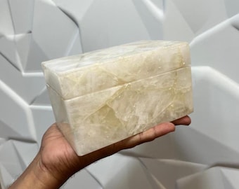 May include: A rectangular box crafted from translucent white and pale yellow stone with a visible crystalline texture and natural veining. The box features a smooth, polished finish and a lid that sits flush with the base, showcasing a marbled appearance.