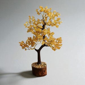 Könnte beinhalten: Eine dekorative Baumskulptur mit braunem Stamm und Ästen sowie gelb-orangen Blättern. Der Baum steht in einem kleinen, runden Holzsockel, der mit kleinen Steinen gefüllt ist. Die Skulptur steht vor einem schlichten, hellgrauen Hintergrund.