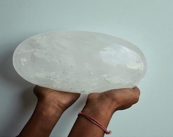 May include: A large, clear quartz crystal in an oval shape. The crystal is held in two hands against a light blue background. The crystal appears to be smooth and translucent, with subtle internal patterns. A pink bracelet is visible on the wrist.