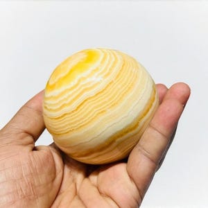 May include: A spherical, polished stone with a banded pattern of yellow and cream hues. The orb is held in a hand against a white background, showcasing the natural variations in the stone's color and texture. The stone's size is approximately 3 inches in diameter.