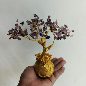 Puede incluir: Un árbol decorativo de piedras preciosas de amatista con una base y ramas doradas. El árbol está adornado con pequeñas piedras de amatista moradas de forma irregular, y las ramas son de alambre dorado. La base es una maceta dorada.