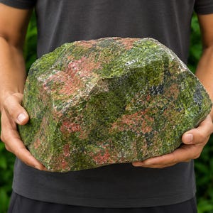 Peut inclure: Un gros morceau de roche brute et non polie présentant une texture tachetée avec des nuances vibrantes de vert forêt, d'olive foncé et des taches de rose saumon. La pierre a une forme angulaire irrégulière avec des dépôts minéraux visibles.