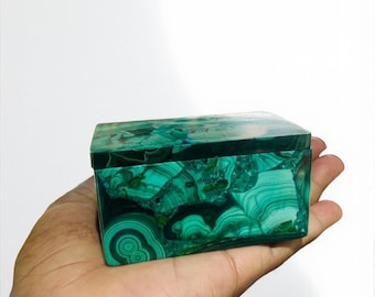 May include: A rectangular box made of malachite, a vibrant green mineral with swirling patterns. The box is held in a hand, showcasing the polished surface and unique natural designs. The box is a decorative item.