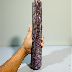 Puede incluir: Un obelisco de cristal cilíndrico y alto con una parte superior puntiaguda. La piedra presenta un patrón moteado de color púrpura oscuro y blanco, con una superficie pulida. El obelisco se sostiene en una mano, mostrando su tamaño y forma. Cristal decorativo.