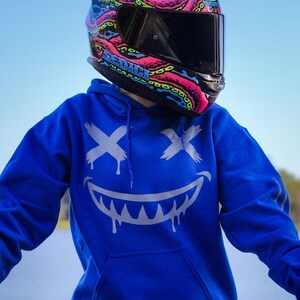 以下が含まれることがあります： 黒いバイザーが付いたオートバイのヘルメット。ピンク、青、緑の触手の鮮やかなデザインで飾られています。ヘルメットには「SEDICE」の文字がプリントされています。その人は、白い笑顔のデザインの青いパーカーを着ています。