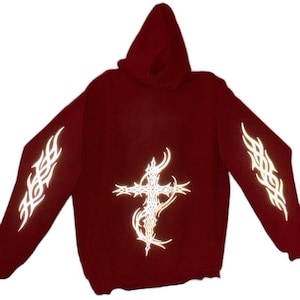 Pu&ograve; includere: Una felpa con cappuccio bordeaux con maniche lunghe. Presenta un design a croce riflettente sul davanti e motivi tribali riflettenti sulle maniche. Il capo &egrave; realizzato in materiale morbido.