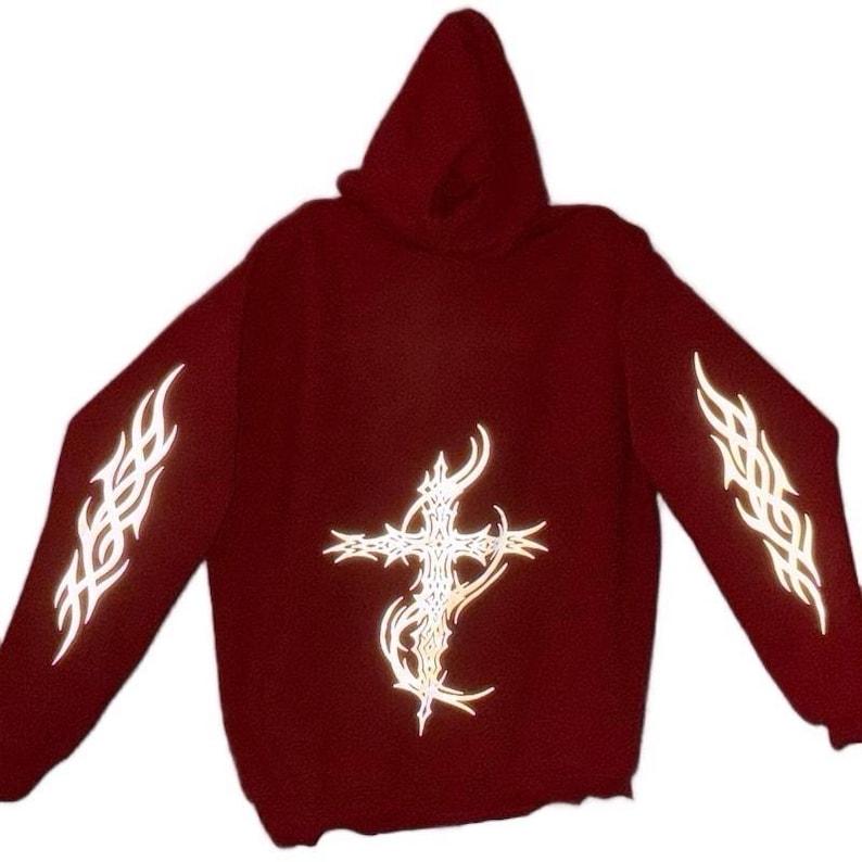 Pu&ograve; includere: Una felpa con cappuccio bordeaux con maniche lunghe. Presenta un design a croce riflettente sul davanti e motivi tribali riflettenti sulle maniche. Il capo &egrave; realizzato in materiale morbido.