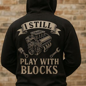 Peut inclure: Sweat à capuche noir avec un dessin d'un moteur, d'un piston et d'une clé. Le texte "I STILL PLAY WITH BLOCKS" est au-dessus et en dessous du dessin du moteur. Le design est de couleur beige clair.