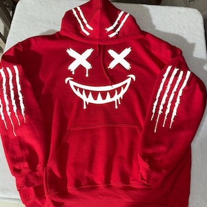 以下が含まれることがあります： 赤いパーカー。白いグラフィックは、滴る牙と十字型の目を持つ笑顔の顔です。白い爪痕が袖とフードを飾っています。パーカーにはフロントポケットとドローストリングフードが付いています。