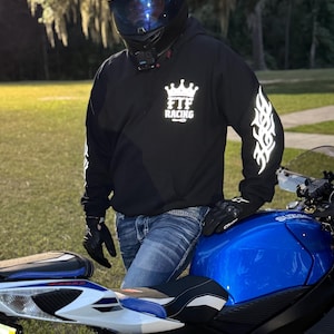 Pu&ograve; includere: Felpa nera con logo "FTF Racing" e design a fiamma sulle maniche. La persona indossa un casco nero con visiera blu, guanti neri e jeans blu. Una moto blu &egrave; in primo piano.