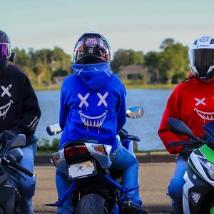以下が含まれることがあります： ヘルメットと笑顔の顔のデザインのパーカーを着た3人のバイク乗り。パーカーは黒、青、赤です。バイクは黒、青、緑です。背景は湖と木々です。