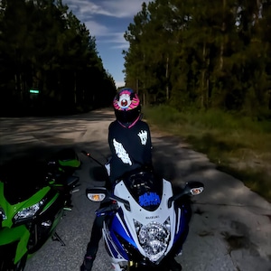 Pu&ograve; includere: Una persona con un casco da moto colorato e una giacca nera &egrave; seduta su una moto Suzuki blu e bianca. Una moto verde &egrave; parcheggiata a sinistra. La scena &egrave; ambientata su una strada buia di notte, con alberi ai lati.