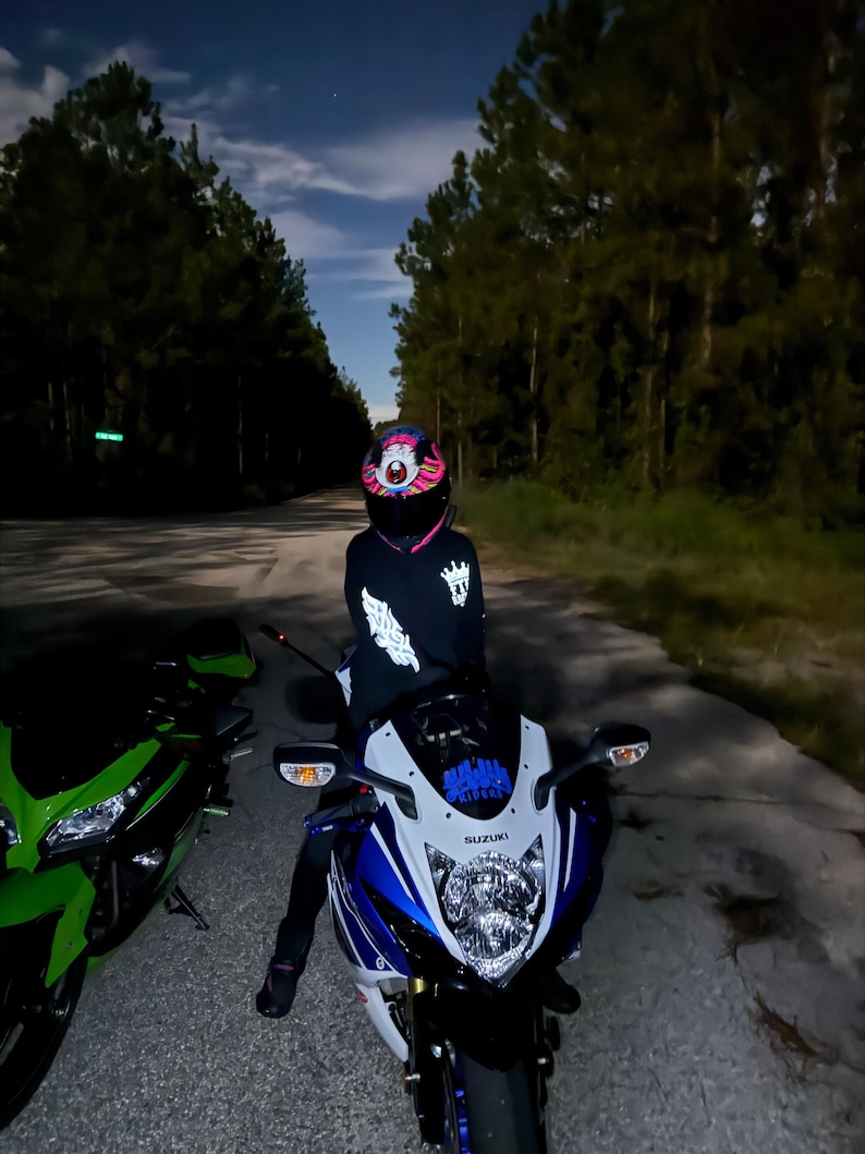Pu&ograve; includere: Una persona con un casco da moto colorato e una giacca nera &egrave; seduta su una moto Suzuki blu e bianca. Una moto verde &egrave; parcheggiata a sinistra. La scena &egrave; ambientata su una strada buia di notte, con alberi ai lati.