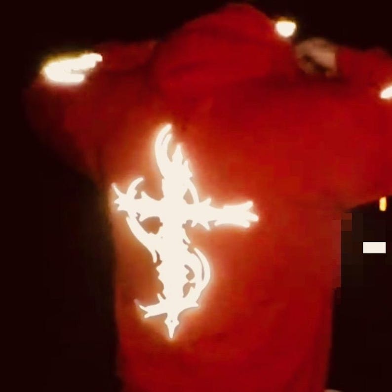 Pu&ograve; includere: Felpa con cappuccio rossa con un design a croce bianca e luminosa sul retro. La felpa ha un cappuccio e maniche lunghe. Il design a croce &egrave; intricato e stilizzato.