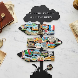 May include: Black metal sign shaped like a directional arrow with the text "OH, THE PLACES WE HAVE BEEN...". The sign is decorated with travel-themed magnets, including landmarks and flags. A travel-themed gift.