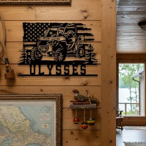 Peut inclure: Décoration murale en métal noir représentant une silhouette de véhicule tout-terrain avec un drapeau américain. Le nom "ULYSSES" est affiché en dessous. L'œuvre est fixée sur un mur en bois.