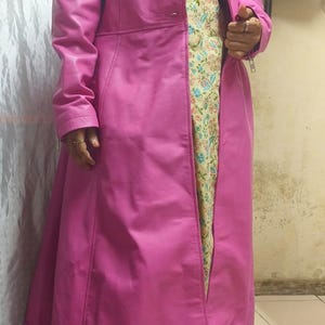 May include: A long, vibrant pink leather coat with a classic lapel collar and a tailored fit. The coat is knee-length and features a single-button closure. The coat is worn over a patterned dress with gold embroidery.