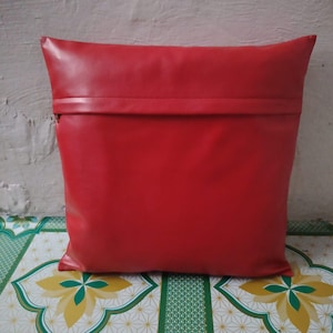 May include: A square, red cushion with a smooth, leather-like texture. The cushion has a folded-over flap at the top, creating a clean, modern design. The cushion is on a patterned surface.