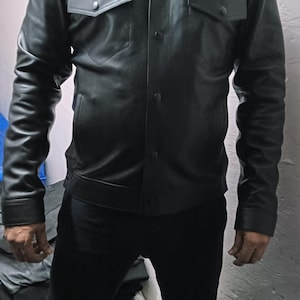 May include: Black leather jacket with a classic collar, two chest pockets, and button closures. The jacket has long sleeves and a tailored fit. The model is wearing black pants. The jacket appears to be new.