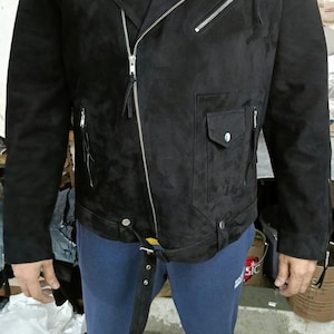 May include: Black suede motorcycle jacket with silver zippers and snaps. The jacket features an asymmetrical zipper closure, a small pocket, and a belt. The model is wearing a yellow shirt underneath.