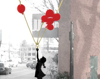 Op de afbeelding: Een gouden ketting met een silhouet van een meisje dat naar rode ballonbedels reikt. De ketting hangt voor een gebouw met een bord waarop "W" staat en een boom op de achtergrond.