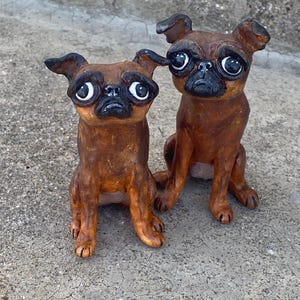 May include: Two brown pug dog figurines with large, expressive eyes and black snouts. The dogs are sitting upright, with their paws visible. The figurines have a glossy finish and are set against a neutral background.