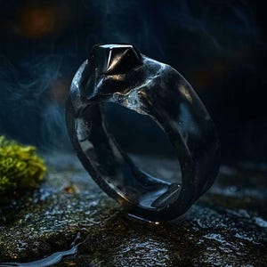 以下が含まれることがあります： 黒い光沢のある指輪。四角くカットされた黒い宝石が特徴です。指輪は暗く質感のある背景に置かれ、左下には緑色の苔が見えます。指輪のバンドは有機的で質感のあるデザインです。