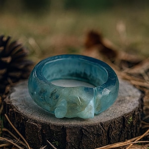 May include: A translucent blue ring with a unique design, resting on a wooden surface. The ring's color shifts from light to darker blue, with a sculpted element. The ring is set against a natural backdrop of wood, pine needles, and foliage.