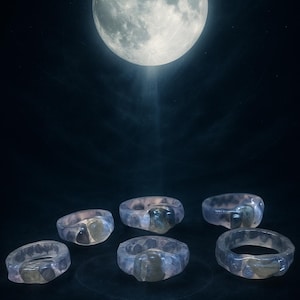 Puede incluir: Seis anillos translúcidos con un centro de piedras preciosas están dispuestos sobre una superficie oscura, iluminados por una brillante luna llena en el cielo nocturno. Los anillos tienen un tono azul claro y un diseño facetado.